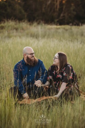 A couple sit on a picnic rug in a long grassy field at sunset