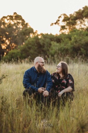 A couple sit on a wooden log in a long grassy field at sunset