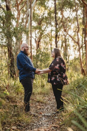 An engaged couple hold hands while standing on a dirt track in bushland