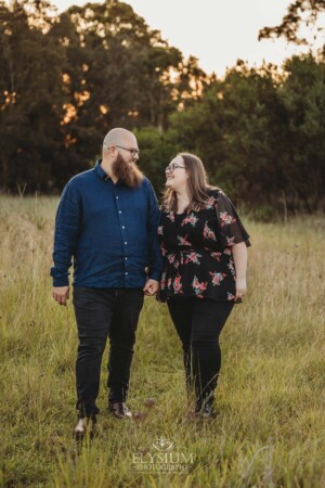 An engaged couple hold hands while walking through long grass at sunset