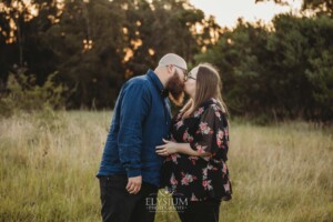 An engaged couple kiss while standing in a grassy field at sunset