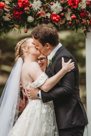 A bride and groom share their first kiss beneath an arbor of bright red florals