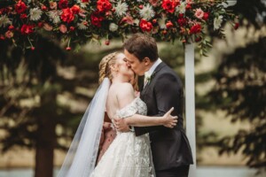 A bride and groom share their first kiss beneath an arbor of bright red florals