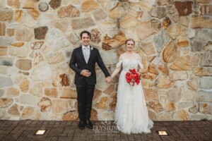 A couple stand against a stone wall and hold hands looking forward