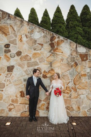 A couple stand against a stone wall and hold hands looking at each other