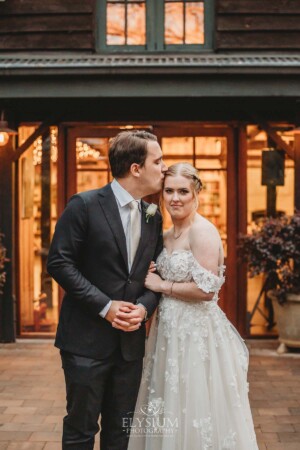 A groom kisses his bride on the cheek as they stand in front of their wedding reception venue