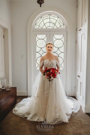 A bride stands in front of a large white door while holding a wedding bouquet of bright red flowers