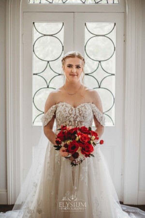 A bride stands in front of a large white door while holding a wedding bouquet of bright red flowers