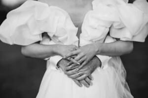 A close up photo of a brides and grooms fingers curled together showing their wedding rings held against the brides dress