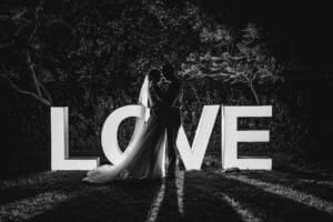 Newlyweds stand in front of an illuminated LOVE sign with a light behind them at night