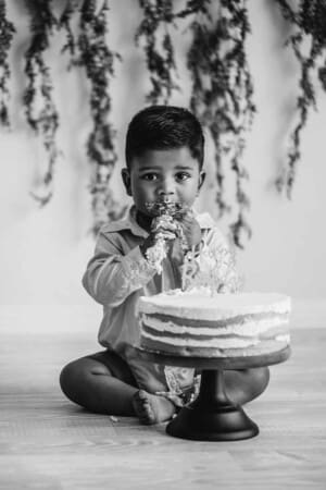 A small boy sitting beside his birthday cake eats it and gets covered in icing