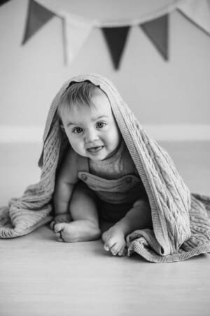 A baby boy sits under a knitted blanket in a natural light Ingleburn studio
