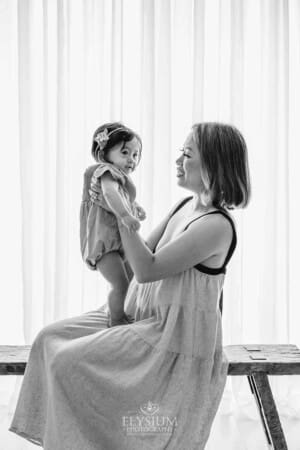 A mother holds her baby in her lap as they sit on a bench in front of a window