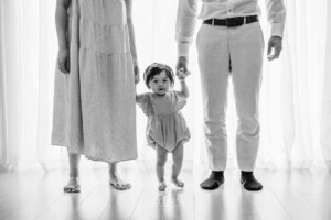 A baby girl stands between her parents holding their hands in front of a large studio window