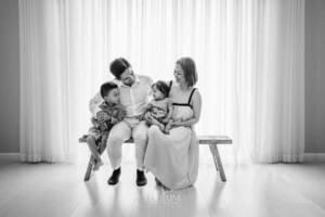 A family sit together on a wooden bench in front of a large bright studio window
