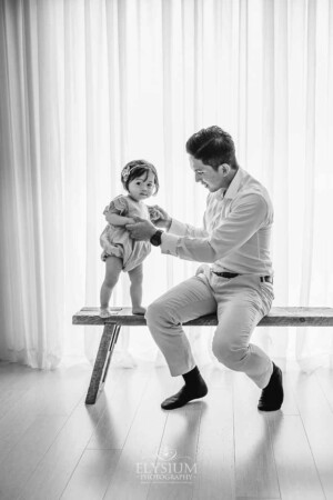 A father holds his baby girls hands as she stands next to him on a timber bench