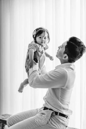 A dad holds his baby girl above him as they sit in front of a large window