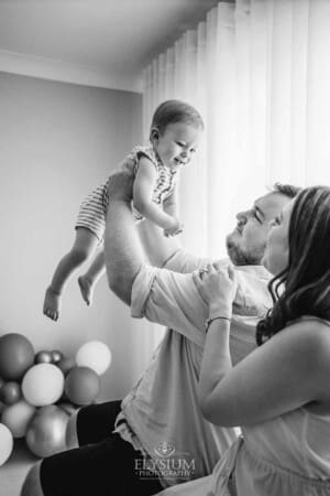 Parents sit in a studio and hold their baby boy above them as they smile for a portrait
