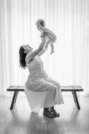 A mother lifts her baby boy above her as she sits on a timber bench in front of a large studio window