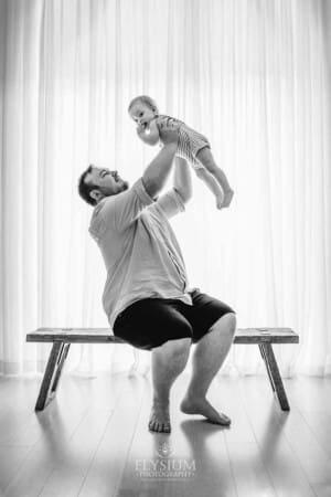 A dad sitting on a wooden bench lifts his baby son above him in front of a large studio window