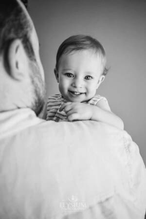 A smiley baby boy peers over his dads shoulder as they stand in a bright studio