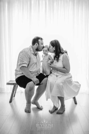 Parents sit with their baby boy on a wooden bench and kiss his face