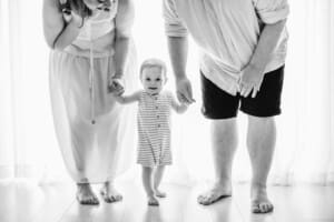 Parents hold their baby boys hands as he stands between them in front of a bright studio window