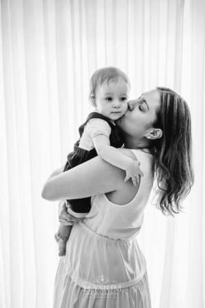 A mother holds her baby boy and kisses his cheek in front of a bright window