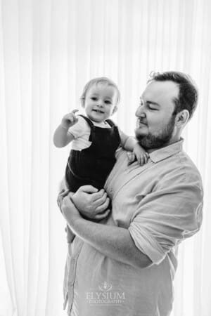 A father cuddles his baby boy as they stand before a bright studio window