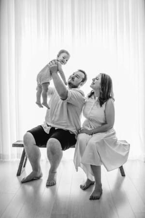Parents sit on a wooden bench in front of a large studio window and lift their smiley baby boy above them