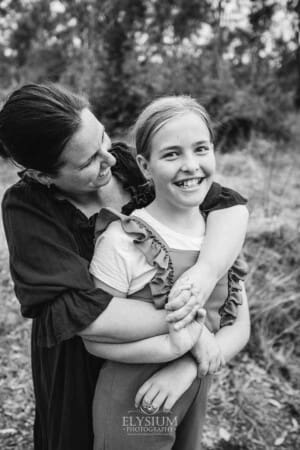 A mother hugs her daughter as they stand in a bushy field