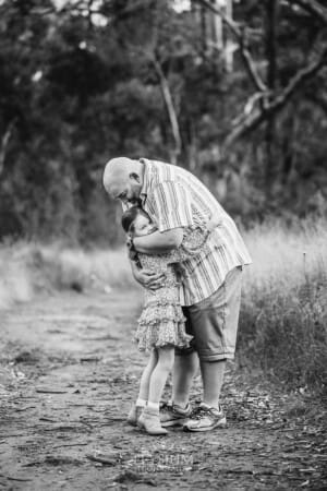 A dad hugs his little girl as they stand on a dirt track in a grass field