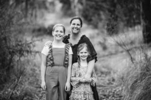 A mother hugs her 2 daughters as they stand on a track surrounded by trees