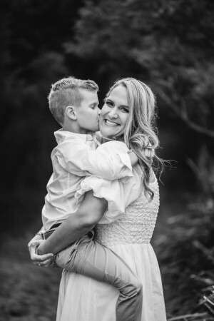 A little boy held by his mother kisses her on the cheek as she looks at the camera