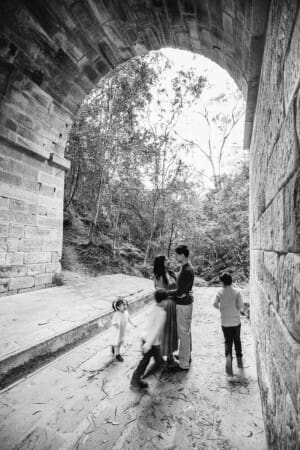 Parents stand under the sandstone arch of Lennox Bridge as their children run around them in a blur