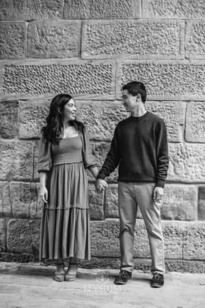 A couple stand against a sandstone wall holding hands and looking at each other