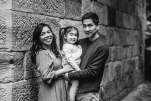 A couple stand next to a stone wall and hold their little girl between them