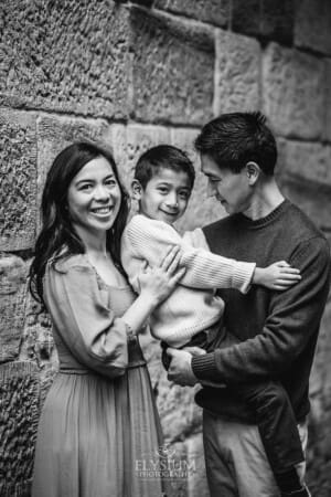 Parents stand beside a stone wall and cuddle their son between them