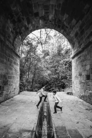 Two little boys jump over a drainage gutter under a sandstone arch at Lennox Bridge