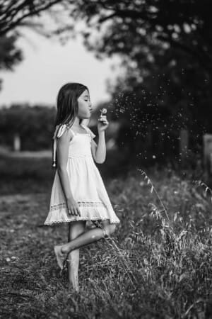 Black and white photograph of a little girl standing in long grass blowing a dandelion