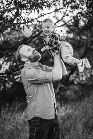A father hoists his baby boy above him as they play and giggle standing in a bushy field