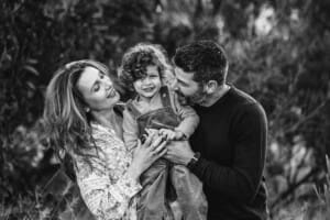 Parents cuddle their baby boy between them as they stand amongst trees with the sun behind them