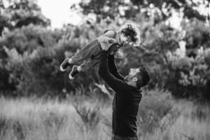 A dad lifts his smiling baby boy above his head as they play in a grassy field