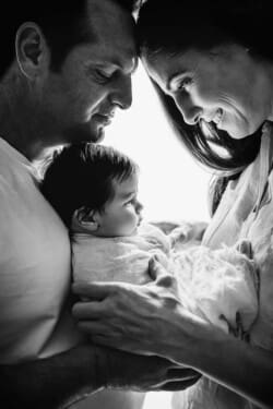 Parents stand close together holding their tiny baby between them with light shining behind them