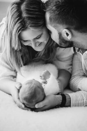 New parents hold their tiny baby between them as the father kisses his wife on the cheek