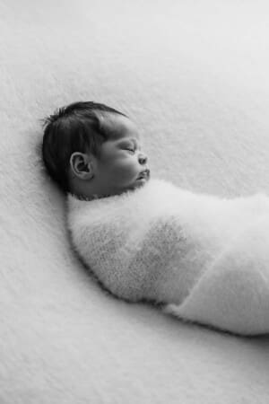 A newborn lays on a white blanket in a soft white wrap sleeping soundly