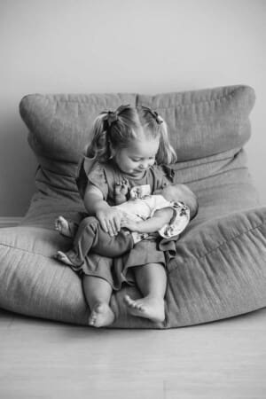 A little girl sittin on a large beanbag holds her newborn baby sister in her lap