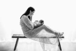 A mother sits on a wooden bench in a white lacey dress holding her newborn baby in her lap