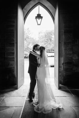 A bride and groom stand in the arched doorway of a church and share a kiss