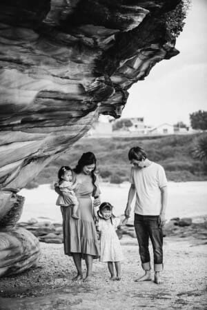 A family stand on a beach next to a sandstone cliff at sunset and hold hands with their little girl